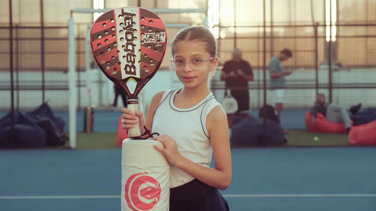 A young girl with glasses holds a paddle racket on an outdoor tennis court during the day.